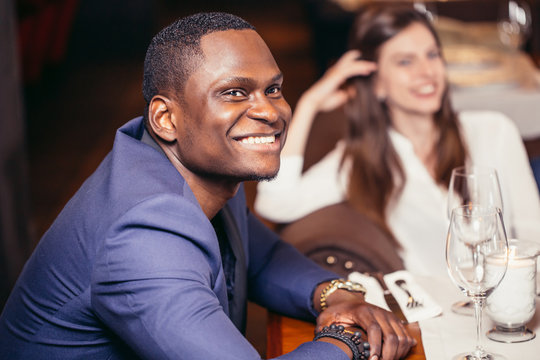 Friendly african guy dressed in expensive jacket looking at camera and smiling while sitting at the table , white woman becide on background