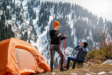 girl with track sticks and a guy with binoculars on a halt near a tourist tent in the mountains examine the surroundings against the background of a snow-covered forest. Leisure. Healthy lifestyle