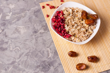 Cooked yummy warm oatmeal and heap of ripe fresh pomegranate seeds and three dried dates in white ceramic bowl on clean bamboo rug on worn gray scratched concrete with copy space