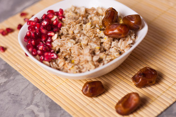 Cooked yummy warm oatmeal and heap of ripe fresh pomegranate seeds and three dried dates in white ceramic bowl on clean bamboo rug on worn gray scratched concrete with copy space