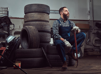 A bearded mechanic dressed in a uniform, sits on old car tires in the garage.