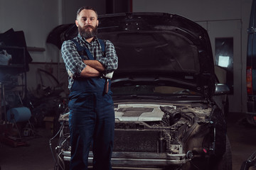 A bearded expert mechanic dressed in a uniform, standing with crossed arms against a car in the...