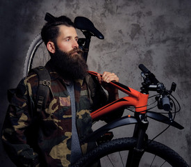 A handsome bearded traveler, dressed in a trendy camouflaged jacket, standing with sports bicycle on a shoulder in a studio, isolated on a dark background.