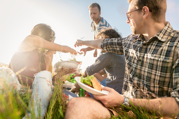 Young people having summer barbecue picnic and eating together sitting in grass