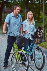 Fototapeta premium An attractive couple of a blonde female and man dressed in casual clothes on a bicycle ride with their cute little spitz in the park.