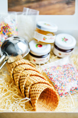 A Kit For Making Ice Cream: Spoon For Ice Cream, Ice Cream Cones, Jam, Topping, Chocolate, Sprinkles, Coconut. In Carton Wooden Natural Background. The Concept Set Surprise, Gift Idea