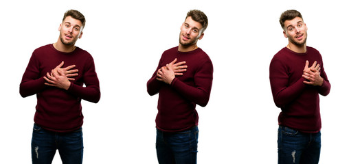 Handsome blond man having charming smile holding hands on heart wanting to show love and sympathy isolated over white background