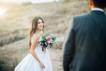 Young bride with wedding bouquet at the hands looking to groom with smile at sunset. Outdoors