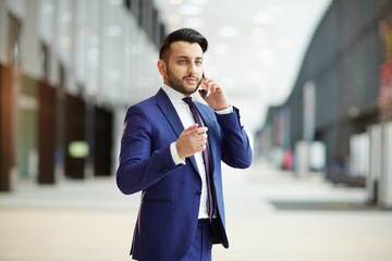 Young confident broker in formalwear talking to one of his clients on smartphone during travel