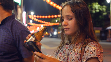 A young girl rests in the evening, drinks beer from a bottle, looks into the phone. HD