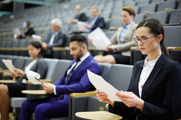 Young serious businesswoman and other people in formalwear reading papers in auditorium or...