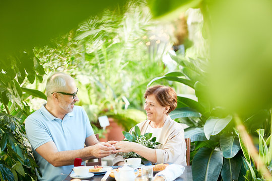 Senior Man Putting Ring On Finger Of His Wife By Romantic Dinner In Orangery Or Home Garden Among Green Plants