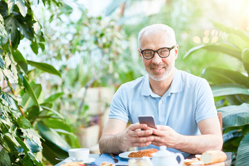 Happy man in eyeglasses looking at camera while texting in smartphone by served table during breakfast