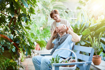 Playful senior woman covering eyes of her husband by hands while both having rest in orangery with...