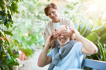 Mature wife covering her husband eyes by hands during their rest in orangery among green plants