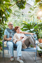 Fototapeta premium Mature couple with glasses of red wine relaxing on bench in orangery of sanatorium or tourist resort