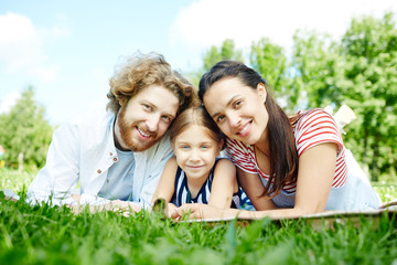 Fototapeta premium Young couple and their daughter looking at camera while relaxing on green lawn in natural environment