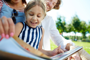 Happy little girl and her parents reading book while spending time in park on summer weekend