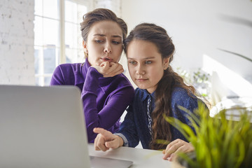 Pretty girl sitting in front of open portable computer at home, pointing finger at screen, showing modern bicycle on web store, mother next to her feeling uncertain and shocked because of high price