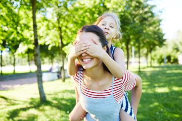 Fototapeta premium Little daughter covering eyes of her mother while sitting on her back and having fun in natural environment