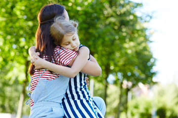 Happy girl sitting on her mother knee while embracing her in park on summer day