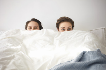 Joy, happiness and fun concept. Picture of playful cheerful European girl and her mother playing hide and seek in bedroom, hiding themselves under white blanket and staring at camera mysteriously