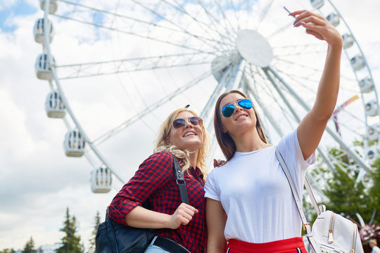Two happy girlfriends in sunglasses and casualwear making selfie on background of enormous ferris wheel in modern theme park