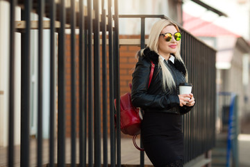 beautiful young girl in a black jacket and glasses on the street with a cup of coffee in hands