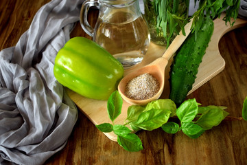 A green pepper, brans in a wooden spoon, basil, tarragon, a cucumber and water in a glass jug. Ingredients for preparing a healthy meal