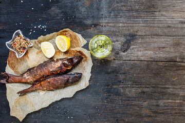 Smoked fish with lemon slices on backing paper on dark wooden smoking background, top view, copy...