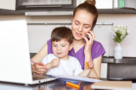 Portrait of young mother works freelance on laptop computer, communicates with someone via smart phone, looks after her little son who doesn`t go to kindergarten. Working mum with male child.