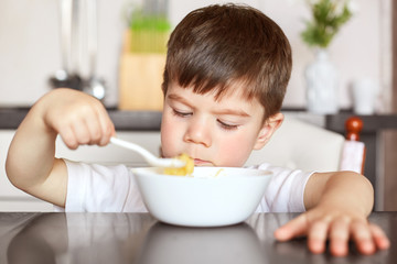 Obraz na płótnie Canvas Healthy eating and children concept. Handsome small child eats with great appetite delicious porridge prepared by mother, holds big spoon and looks at bowl, sits against domestic kitchen interior