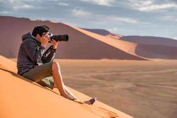 Young Asian man traveler and photographer sitting on sand dune taking photo of sunset in Namib...
