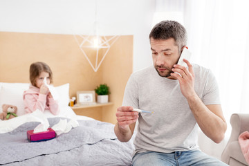 Terrible sickness. Frightened dark-haired daddy holding a thermometer and talking on the phone with the doctor