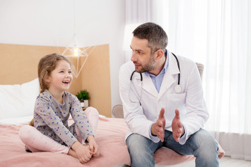 So happy. Adorable joyful girl sitting on bed and talking with the doctor