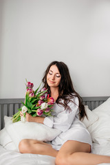 beautiful young brunette girl in white shirt with bouquet of tulips on bed at home