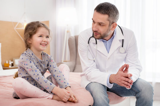 Delighted. Charming joyful girl sitting on bed and talking with the doctor