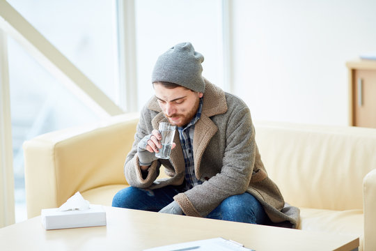 Young homeless man in shabby clothes sitting on sofa in social support center and drinking water