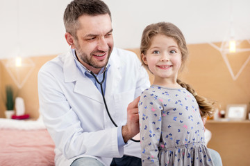 Feeling good. Delighted skilled doctor examining his patient and listening to the girls heartbeat