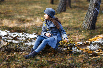 A girl in grey coat and a cute gray hat in the forest in early spring with a willow branch of twigs smelling twigs. A girl outside after rain at the sunset