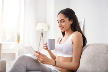 Obraz premium Cozy reading. Pleasant young woman sitting on the sofa and reading from tablet while drinking tea and smiling