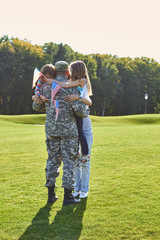 Soldier reunite with his family outdoors. Back side view USA soldier in camouflage is hugging with family.