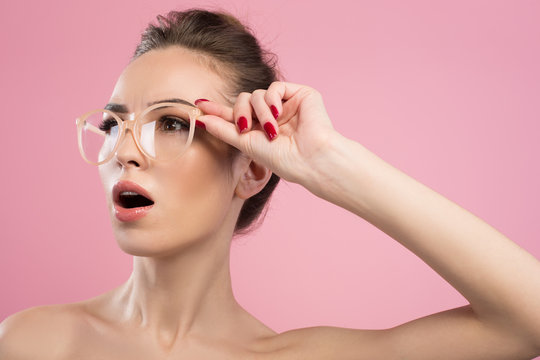 Surprise. Young woman in glasses is looking aside having ones eyes wide open in amazement. Isolated on pink background