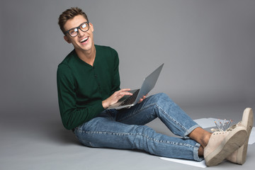 Portrait of smart guy sitting with computer on knees and rejoicing. He is wearing pair of spectacles