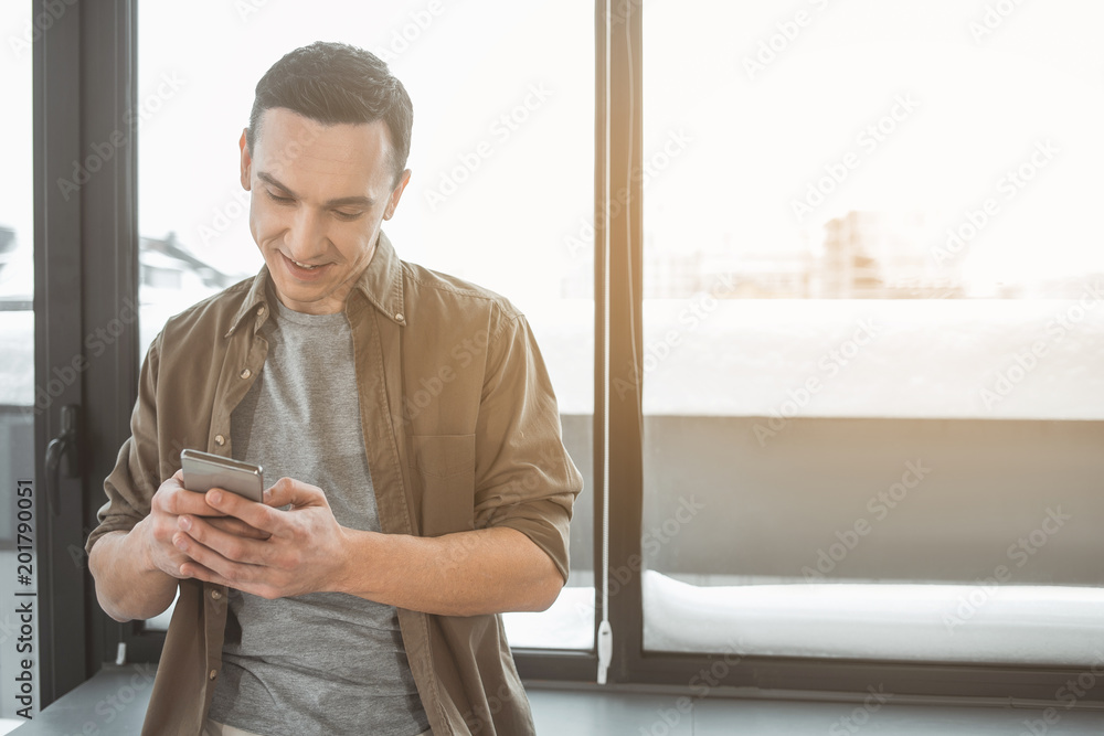 Poster portrait of cheerful man typing in smartphone while standing near window. job with technology concep