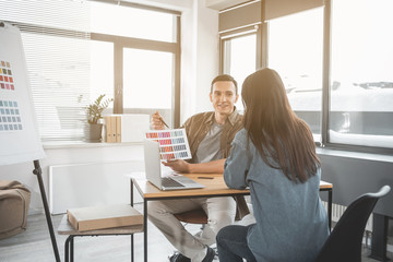 Portrait of beaming stylist of interior proposing different colors for walls. He talking with female client at desk. Creative occupation concept