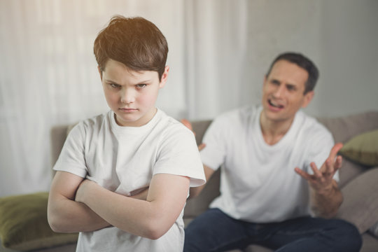Portrait of abused boy standing with crossed hands in room. Angry father is yelling at kid and gesturing with frustration on background