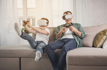 Joyful father and son are playing video game and laughing. They are using virtual reality headsets while sitting on sofa at home