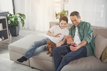 Joyful man and boy speaking online while waving hands to smartphone camera. They are relaxing on sofa with dachshund pet and laughing