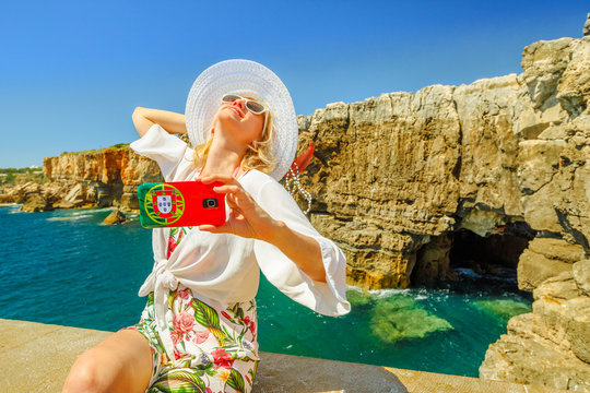 Woman takes photo of Boca do Inferno, a cliff formation, Cascais, Atlantic Ocean, Portugal.Female tourist takes pictures by mobile phone with Portugal flag of Hell's Mouth a touristic sightseeing spot
